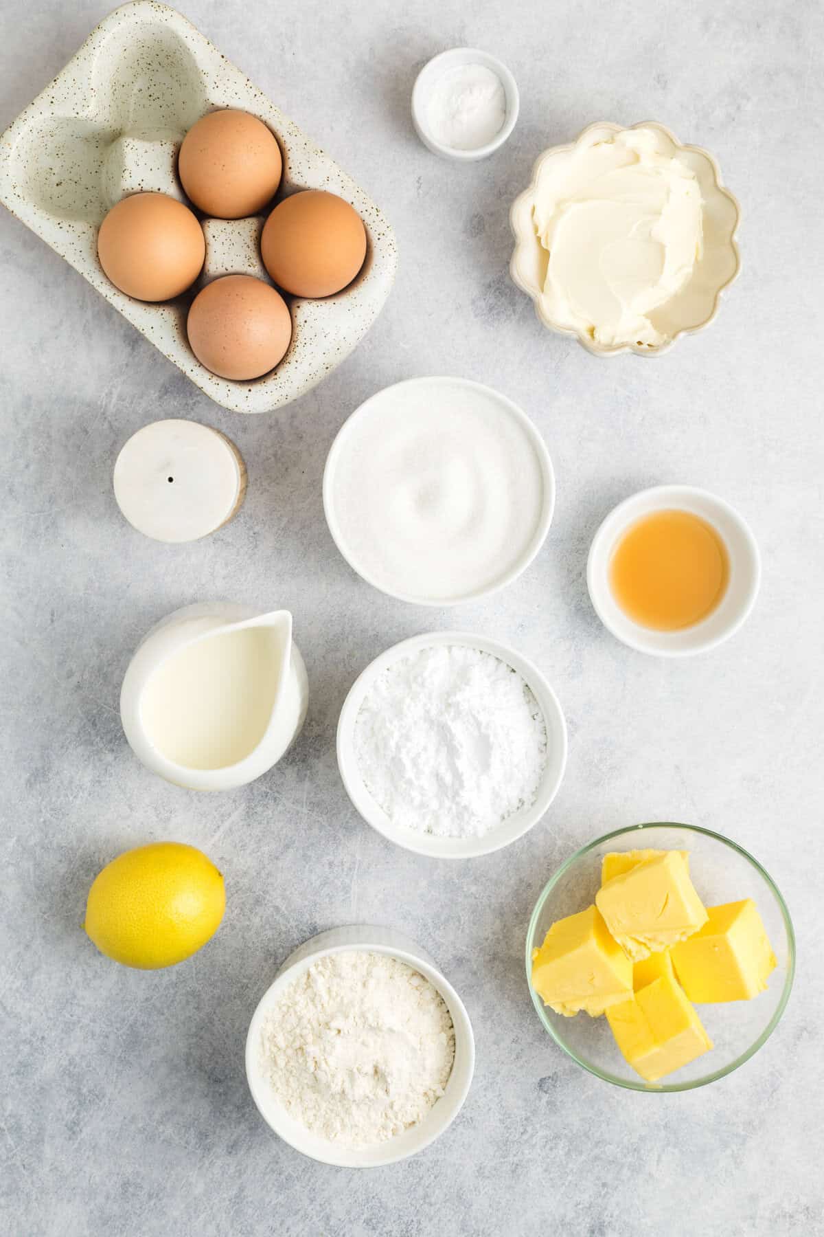 Assorted baking ingredients on a light surface, including eggs, butter, flour, sugar, milk, lemon, vanilla, baking powder, and softened cream cheese arranged in small bowls.