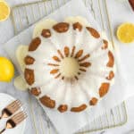 A lemon bundt cake with white glaze sits on parchment atop a cooling rack, surrounded by fresh lemons. Two forks rest on a white plate nearby.