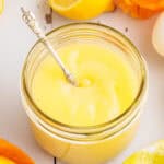 A glass jar filled with creamy yellow lemon curd, with a silver spoon inside. Slices of lemon and a white egg are nearby on a white wooden surface.