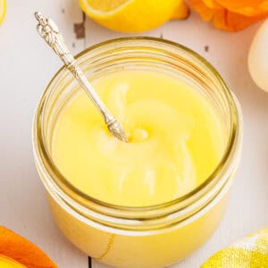 A glass jar filled with creamy yellow lemon curd, with a silver spoon inside. Slices of lemon and a white egg are nearby on a white wooden surface.