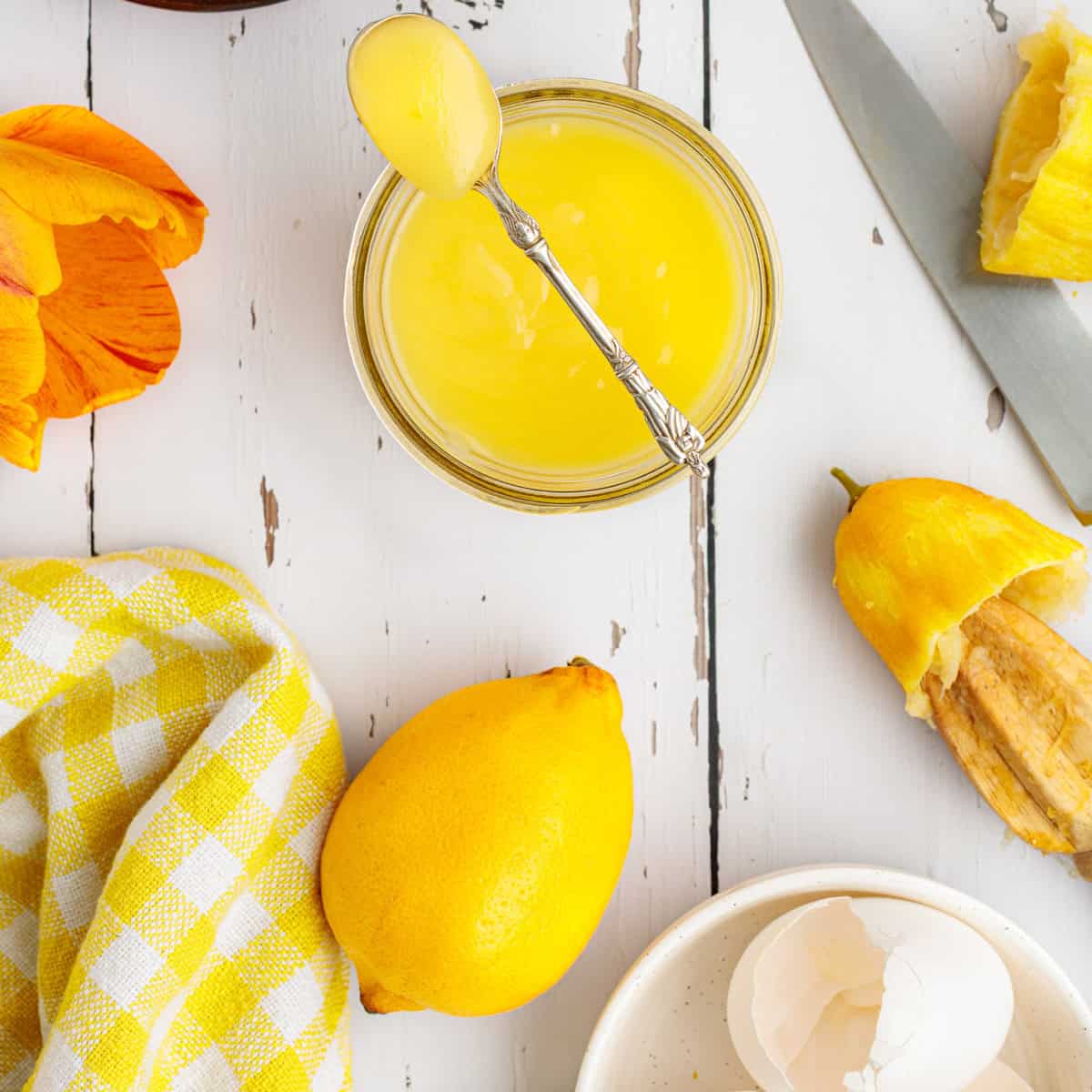 A jar of lemon curd with a spoon, a whole lemon, lemon peel, egg shells, a yellow checkered cloth, a knife, and part of an orange flower are artfully arranged on a white wooden table.
