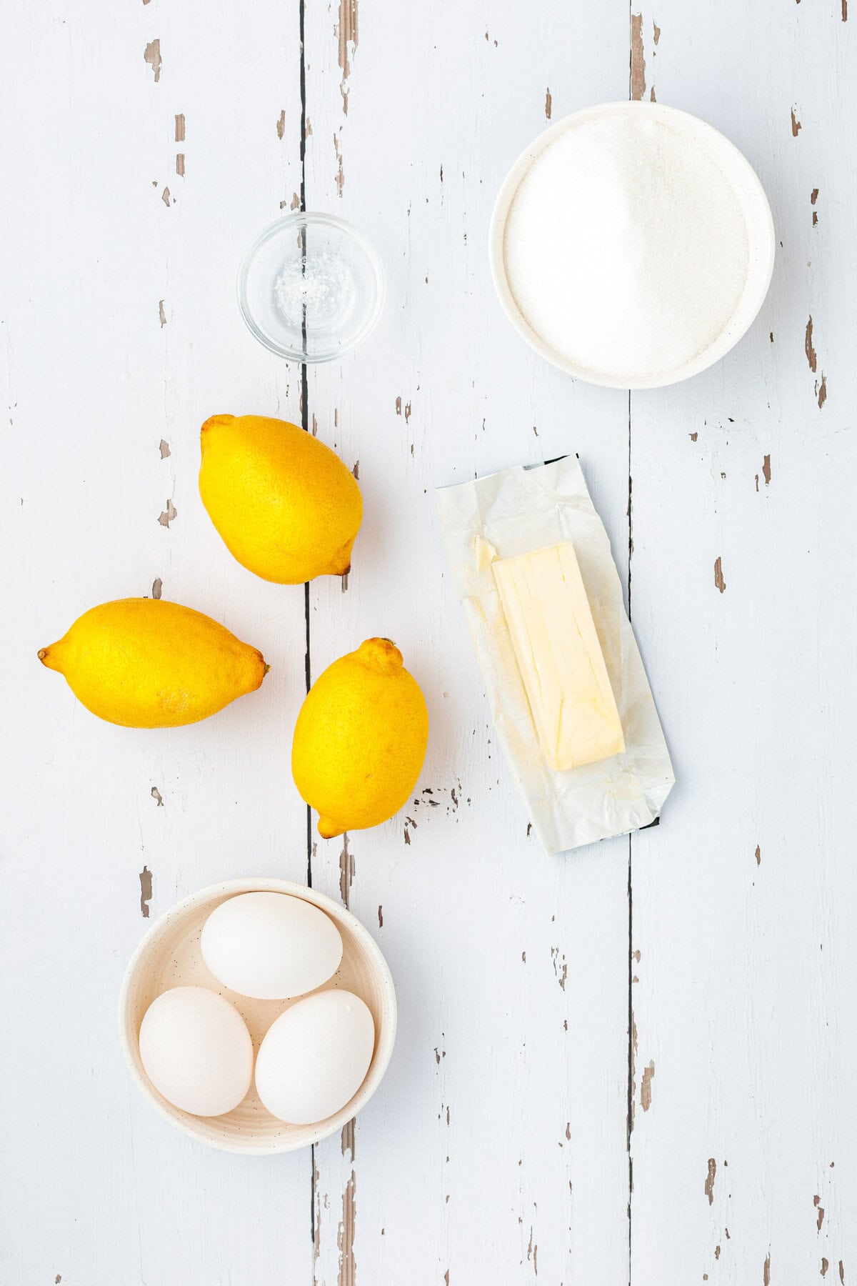 Three lemons, three eggs in a bowl, a stick of butter with wrapper, a bowl of sugar, and a small glass dish, all arranged on a white rustic wooden surface.