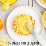 A toasted English muffin spread with lemon curd on a white plate, with a jar of lemon curd and a spoon beside it. Text reads: Homemade Small Batch Lemon Curd. Visit www.theurbankitten.com..