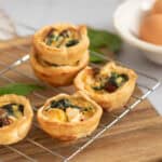 Four mini quiches with spinach and sun-dried tomatoes sit on a cooling rack atop a wooden board, with fresh eggs and green leaves blurred in the background.