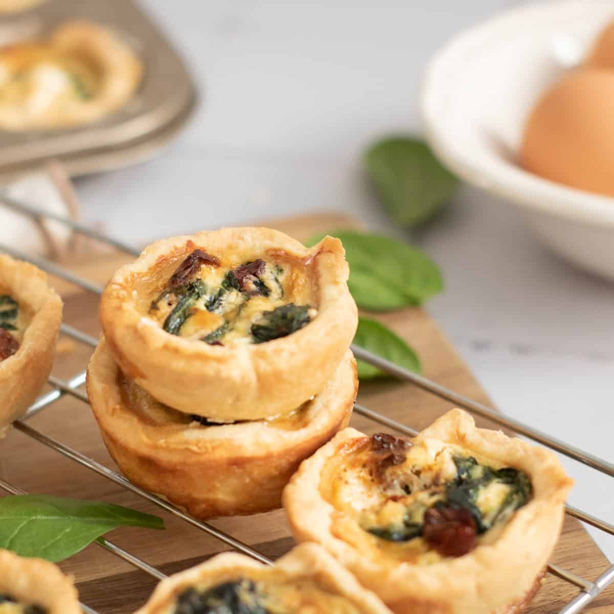 Mini quiches with spinach and sun-dried tomatoes stacked on a cooling rack, with a muffin tin and a bowl of eggs in the blurred background.