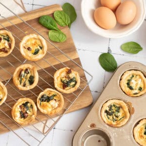 Mini spinach quiches on a cooling rack and in a muffin tin, with fresh spinach leaves scattered and a bowl of eggs nearby on a white surface.