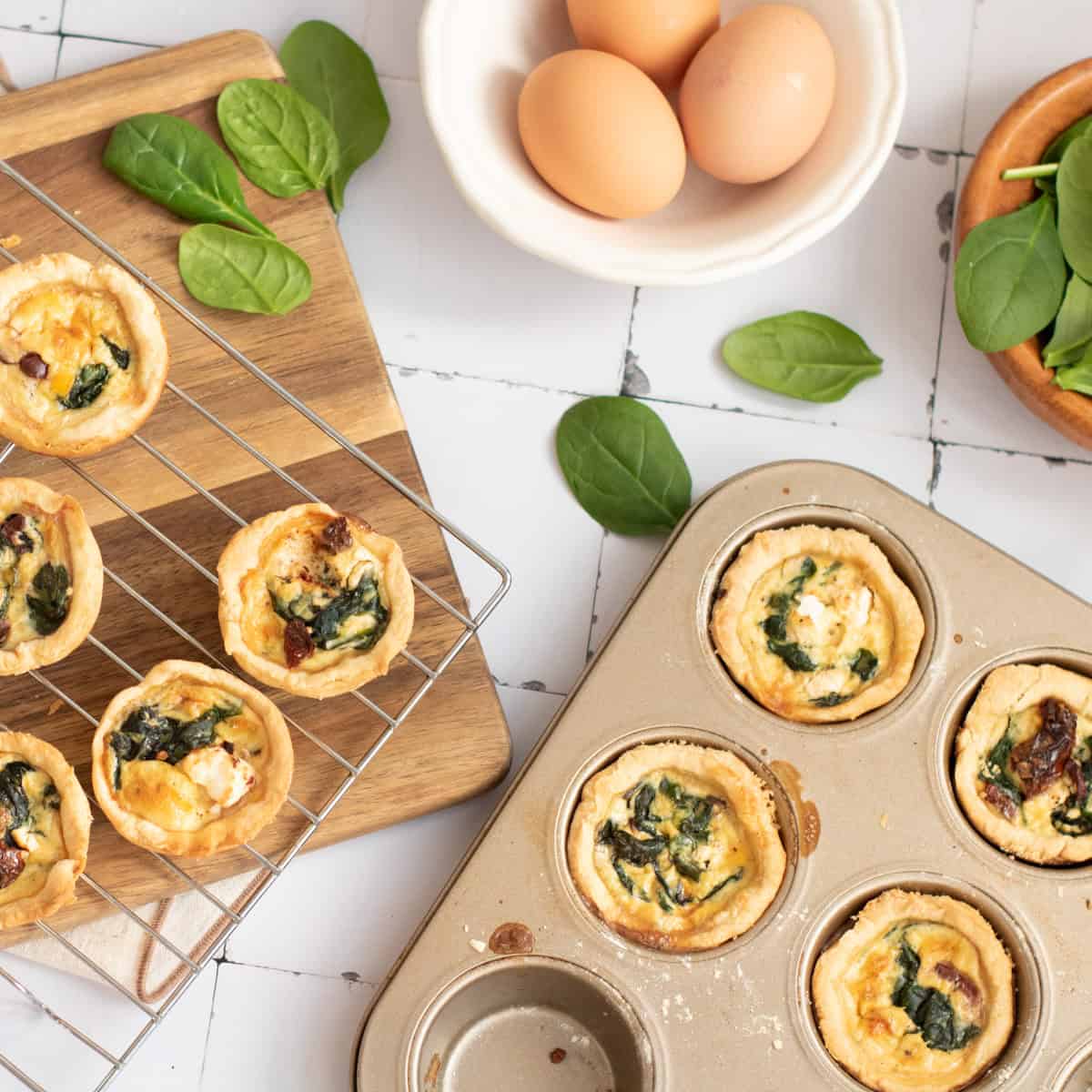 Mini spinach and cheese quiches in a muffin pan and cooling rack, surrounded by fresh spinach leaves and three eggs in a bowl on a white tiled surface.