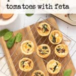 Mini tarts with spinach, sun-dried tomatoes, and feta on a cooling rack, with fresh spinach leaves scattered nearby. A wooden board and white table surface are underneath.