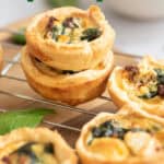 Mini spinach quiches stacked on a cooling rack, with golden crusts and visible spinach and cheese filling. Text on image reads mini party size spinach quiche.