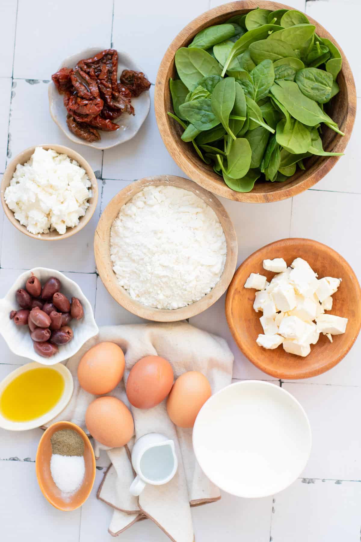 A flat lay of ingredients including fresh spinach, eggs, flour, feta cheese, goat cheese, sun-dried tomatoes, Kalamata olives, olive oil, cream, salt, and pepper, all arranged in bowls on a white surface.