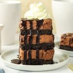 A tall slice of layered chocolate cheesecake with whipped cream on top and chocolate sauce drizzled over it, served on a white plate.