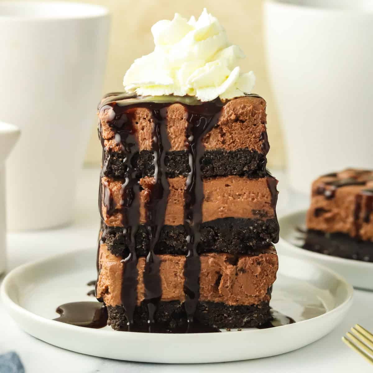 A tall slice of layered chocolate cheesecake with whipped cream on top and chocolate sauce drizzled over it, served on a white plate.