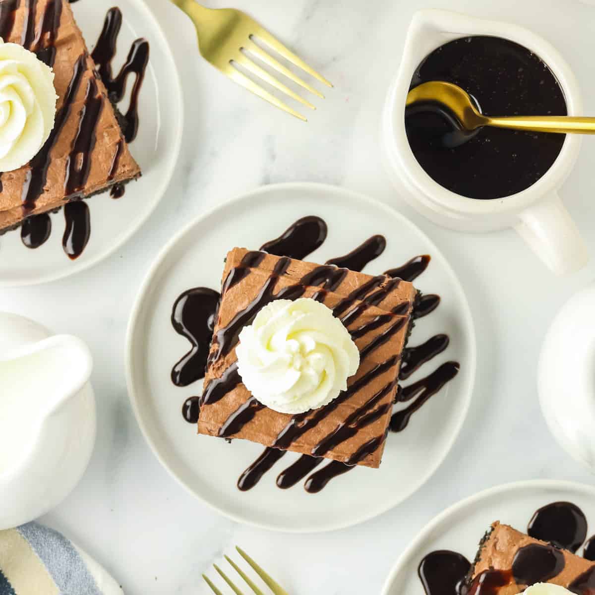 A plate with a square slice of chocolate cake topped with whipped cream and chocolate drizzle, surrounded by golden forks, a pitcher of chocolate sauce, and white dishes on a white surface.
