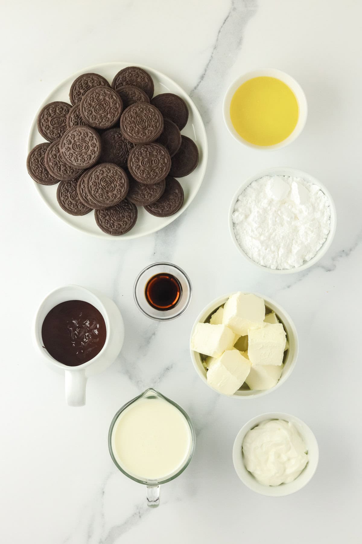 A marble surface with bowls of chocolate sandwich cookies, melted butter, powdered sugar, cream cheese, vanilla extract, whipped cream, chocolate sauce, and sour cream arranged neatly.