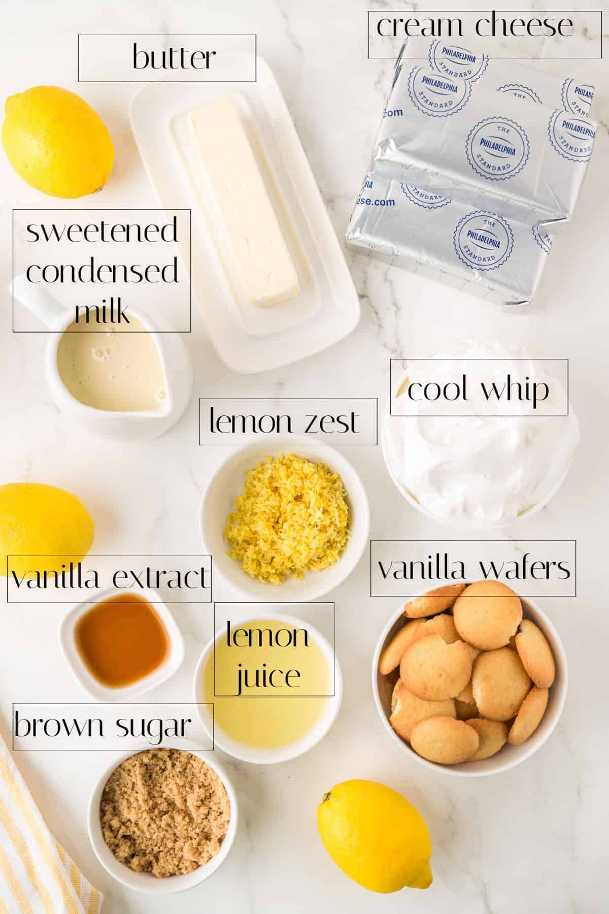 Ingredients for a lemon dessert are arranged on a white surface, including butter, cream cheese, sweetened condensed milk, lemon zest, cool whip, vanilla wafers, vanilla extract, lemon juice, brown sugar, and lemons.