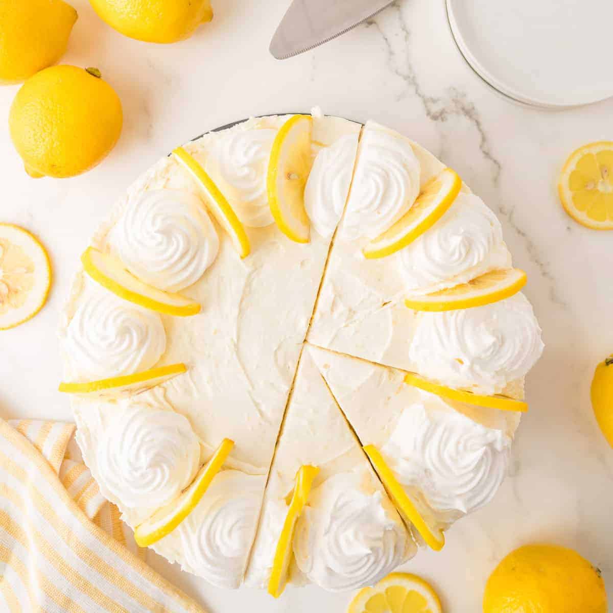 Top view of a lemon cake with whipped cream swirls and lemon slices on top, partially sliced, surrounded by whole and sliced lemons on a white marble surface.