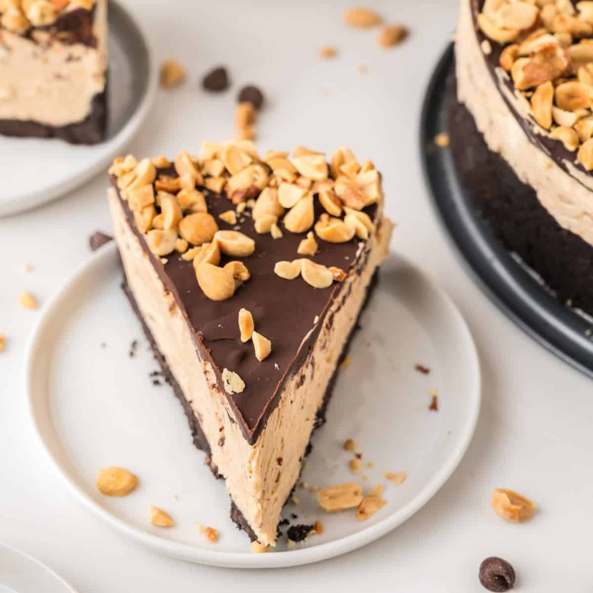 A slice of peanut butter cheesecake with a chocolate topping and chopped peanuts sits on a white plate. The full cheesecake and scattered peanuts are visible in the background.
