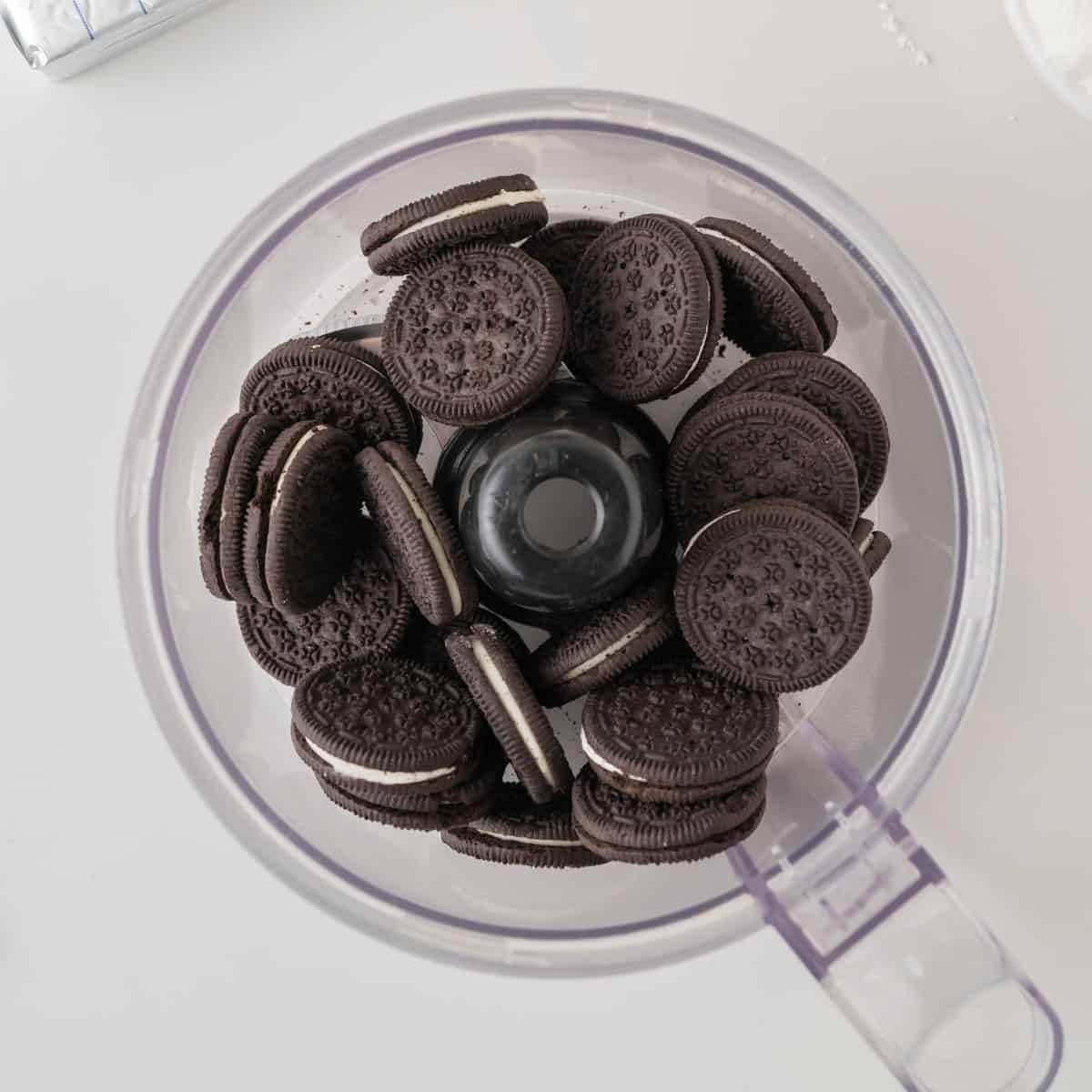Several chocolate sandwich cookies with white filling are placed inside a clear food processor bowl, viewed from above on a white surface.
