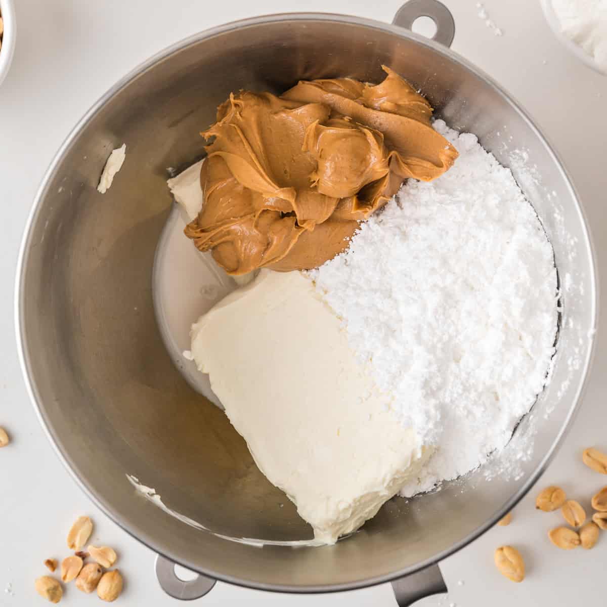 A metal mixing bowl with cream cheese, peanut butter, and powdered sugar, ready to be mixed. A few peanuts are scattered nearby on the white surface.