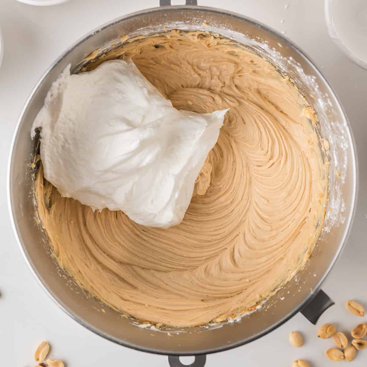 A metal bowl contains light brown batter with a mound of fluffy white whipped topping on one side. Nearby, there are a few peanuts and white bowls, all set on a white surface.