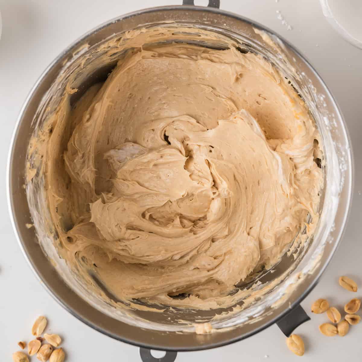 A metal mixing bowl filled with creamy, light brown peanut butter frosting. Shelled peanuts are scattered nearby on a white surface.