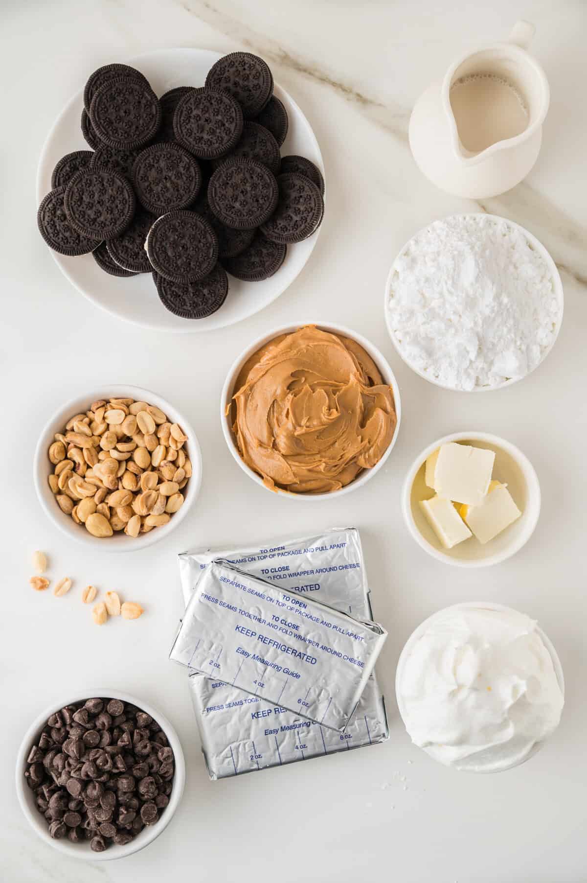 A variety of dessert ingredients on a white surface, including chocolate sandwich cookies, peanuts, chocolate chips, peanut butter, cream cheese, whipped topping, butter, powdered sugar, and a small pitcher of milk.