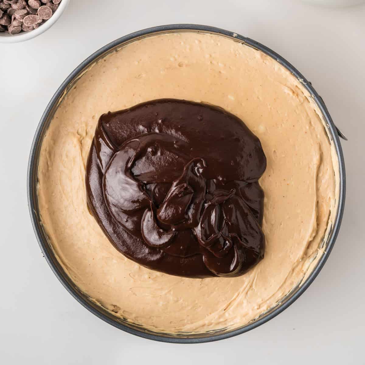A round pan filled with creamy cheesecake batter topped with a large swirl of glossy chocolate ganache, ready to be spread.