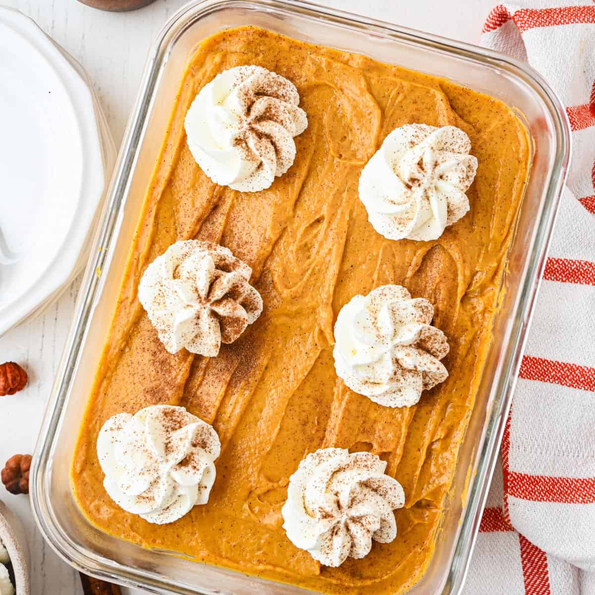 A glass dish filled with pumpkin dessert topped with six swirls of whipped cream, dusted with cinnamon. A red-striped towel and plates are nearby.