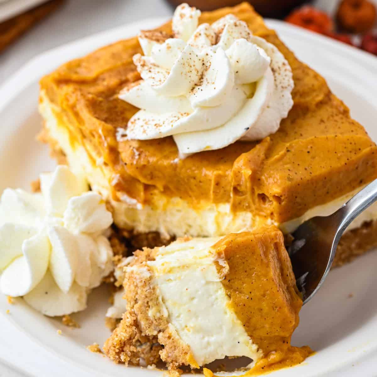 A slice of pumpkin cheesecake with a graham cracker crust, topped with whipped cream and a sprinkle of cinnamon. A fork holds a bite taken from the creamy dessert on a white plate.