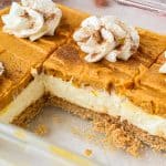 A glass dish of no-bake pumpkin cheesecake sits on the table, showcasing a crumbly crust, creamy middle layer, pumpkin top layer, and whipped cream dollops sprinkled with cinnamon. One large rectangular piece has been cut out and removed.