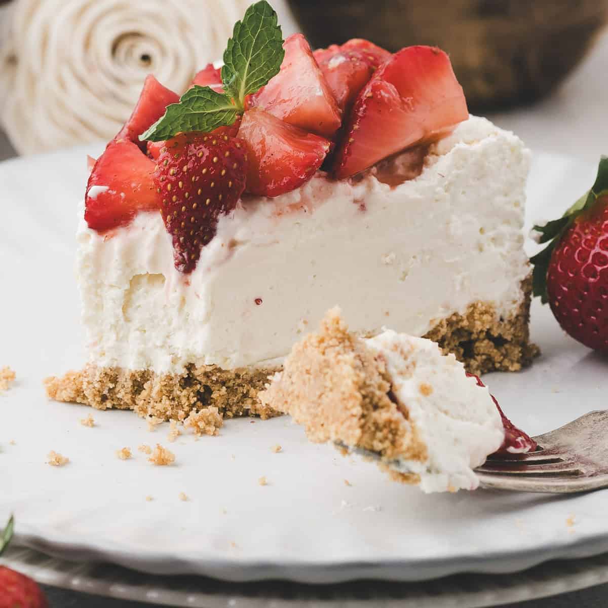 A slice of creamy cheesecake with a graham cracker crust, topped with fresh strawberries and a mint leaf, sits on a white plate with a forkful taken out.