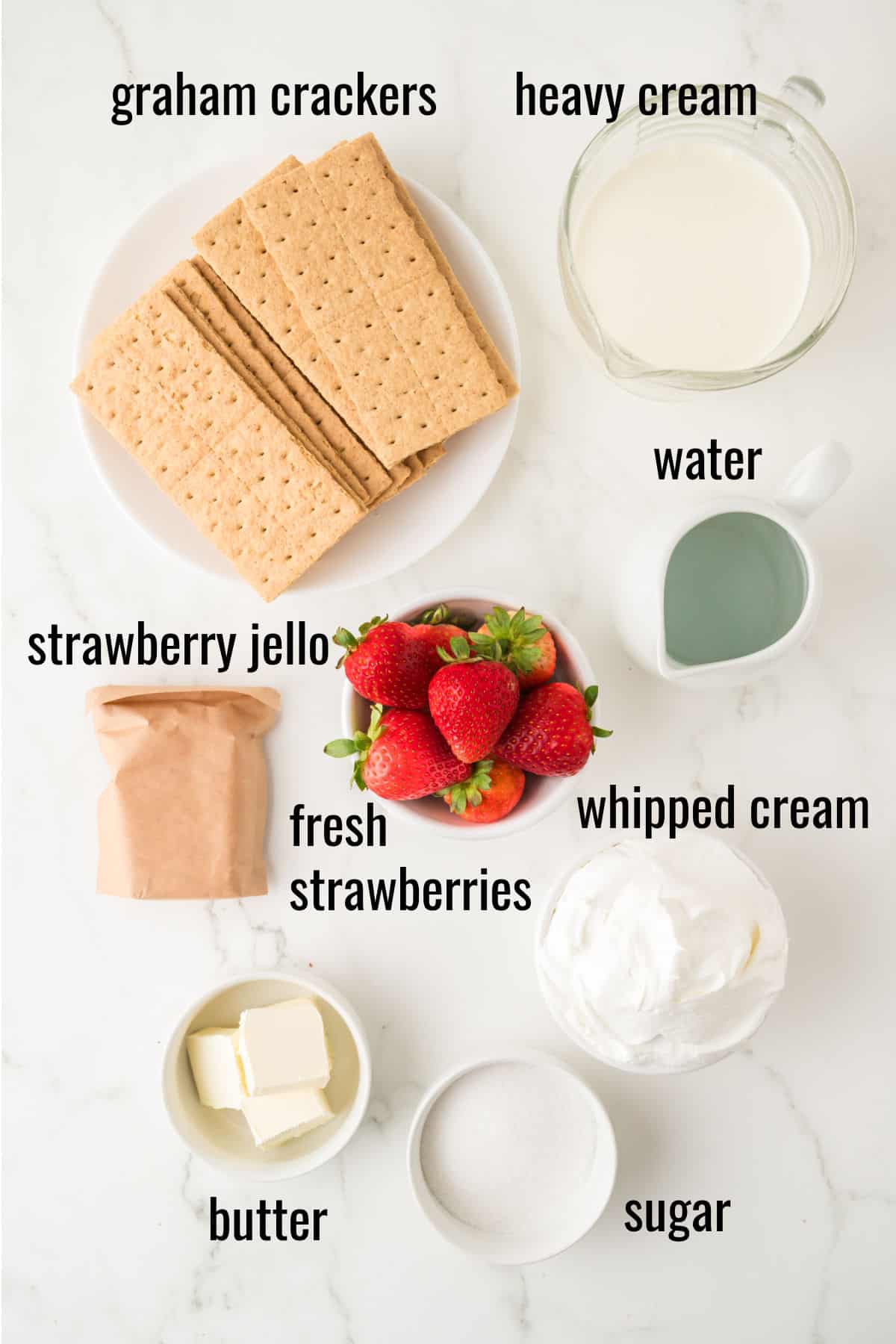 Top view of ingredients on a white surface: graham crackers, heavy cream, water, strawberry jello packet, fresh strawberries, whipped cream, butter, and sugar, each labeled with text.