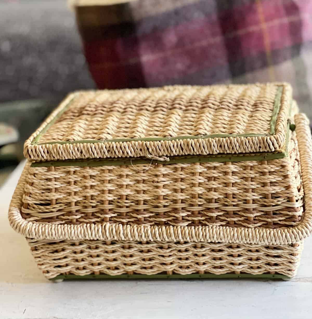 A rectangular woven wicker basket with a lid sits on a white surface. The lid is slightly askew, revealing green trim. In the blurred background, a plaid blanket is visible.