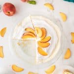 A peach cream pie with a slice missing, topped with peach slices on whipped cream. Peach slices and a whole peach are scattered around on a white marble surface.
