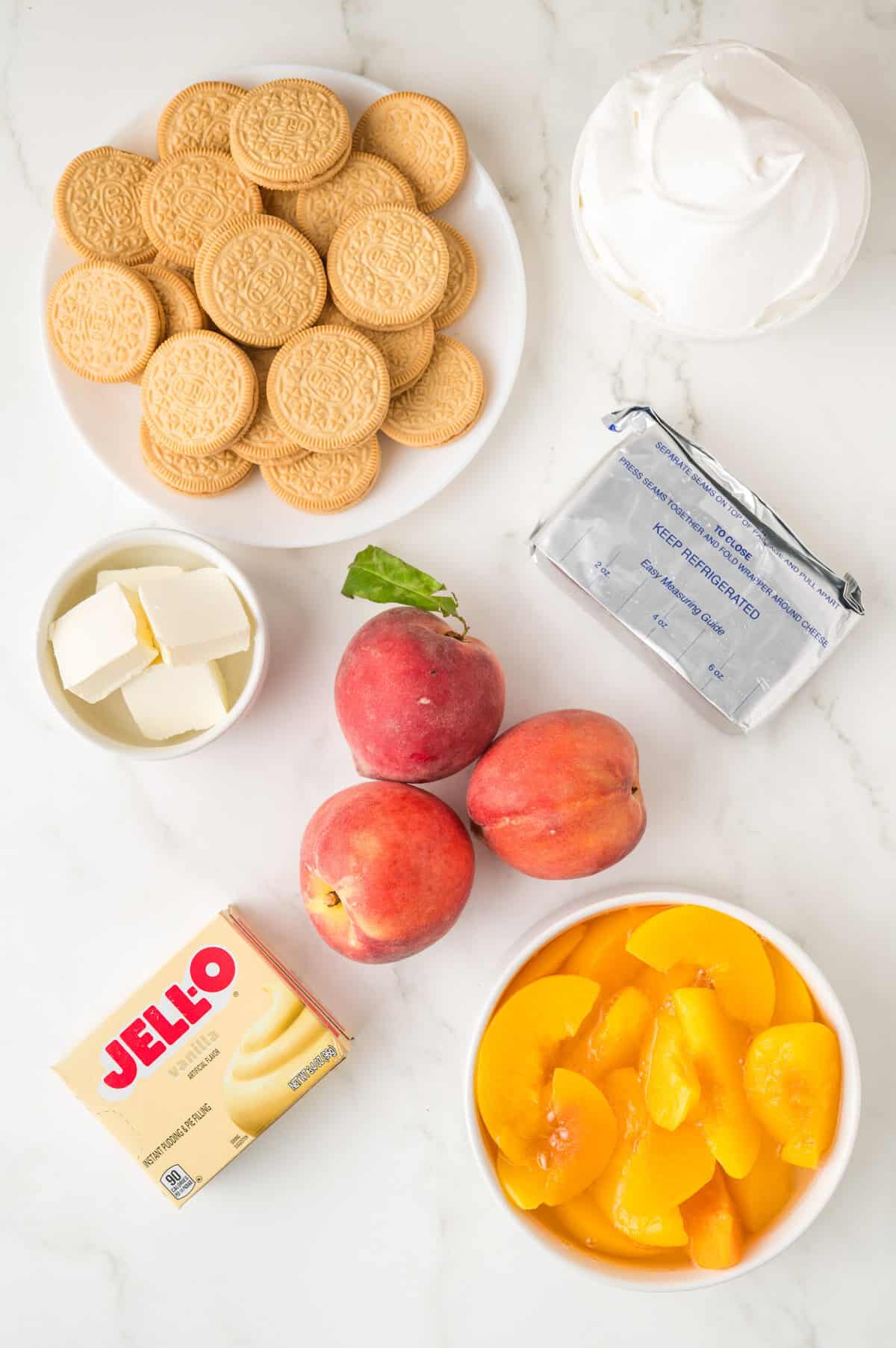 Ingredients on a white surface: a plate of golden sandwich cookies, a bowl of whipped topping, cream cheese, butter, three fresh peaches, a bowl of sliced peaches, and a box of Jell-O vanilla pudding mix.