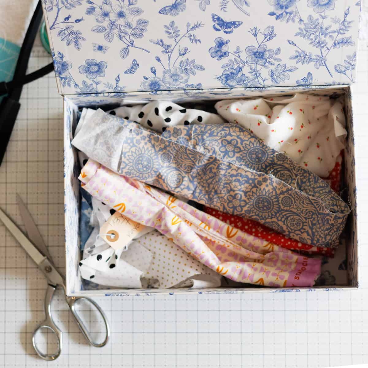 A decorative box with a blue floral pattern holds various folded fabrics in different colors and patterns. A pair of silver scissors lies next to the box on a grid-patterned surface.
