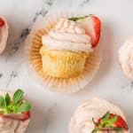 A vanilla cupcake with pink frosting and a sliced strawberry on top, partially unwrapped from its paper liner, sits on a white marble surface surrounded by similar cupcakes.