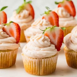 Vanilla cupcakes topped with swirls of light pink frosting and garnished with fresh strawberry halves, arranged on a white surface.