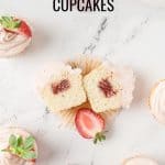 Overhead view of strawberry filled cupcake cut in half, revealing the jam center. Surrounded by frosted cupcakes and fresh strawberries on white marble. Text reads: Strawberry Jam Filled Cupcakes by theurbankitchen.com.