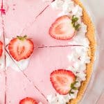 A pink pie with a graham cracker crust, topped with whipped cream and halved strawberries, sliced into pieces on a glass pie plate.