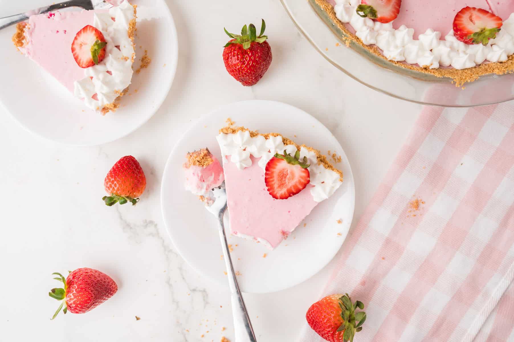 A slice of pink strawberry cream pie with whipped topping and a fresh strawberry, on a white plate with a fork. Whole strawberries and a pie dish are nearby on a marble surface with a pink striped napkin.