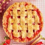 A golden-brown lattice-top strawberry rhubarb pie sits on a cooling rack, with sugar sprinkled on the crust. Fresh strawberries and rhubarb are placed nearby on a pink checkered surface.