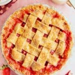 A golden-brown lattice-topped strawberry rhubarb pie sits on a pink surface, with strawberries, rhubarb stalks, a cloth napkin, a pitcher of cream, and stacked plates with forks nearby. The glossy red filling peeks through the crust.