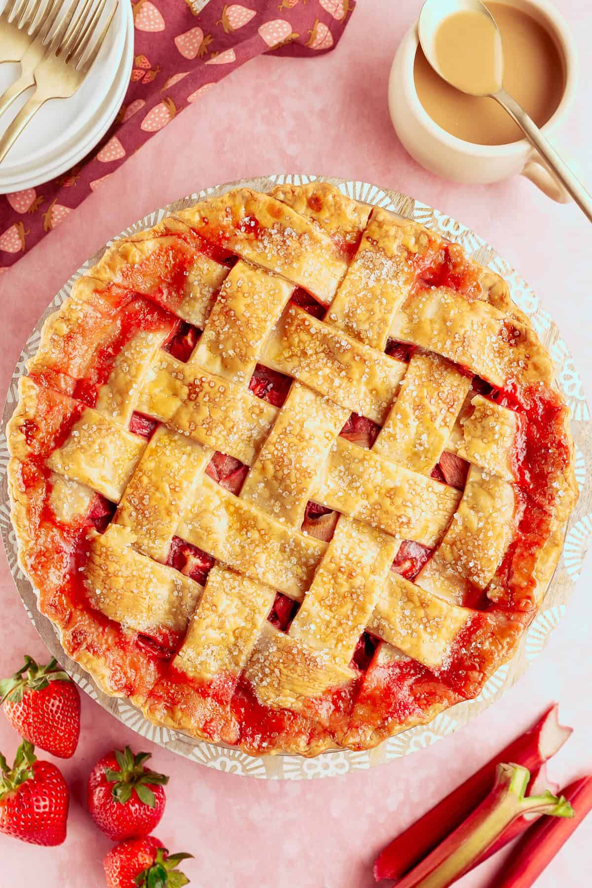 A golden lattice-topped pie filled with strawberry and rhubarb sits on a pink surface, surrounded by fresh strawberries, rhubarb stalks, plates, utensils, and a jug of cream.