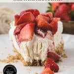 A slice of vanilla cheesecake topped with fresh strawberries sits on a white plate with a crumb crust, surrounded by more strawberries. Text on the image describes the dessert.