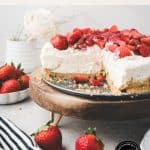 A sliced no-bake vanilla cheesecake topped with fresh strawberries sits on a wooden board. Whole strawberries and a white rose are in the background, enhancing the inviting presentation of this no-bake vanilla cheesecake. Text reads: “no-bake desserts Vanilla Cheesecake.”.