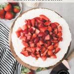 A creamy no-bake vanilla cheesecake topped with chopped fresh strawberries sits on a wooden board, surrounded by whole strawberries and a striped towel. Text over the image reads, “Vanilla Cheesecake with fresh strawberries.”.