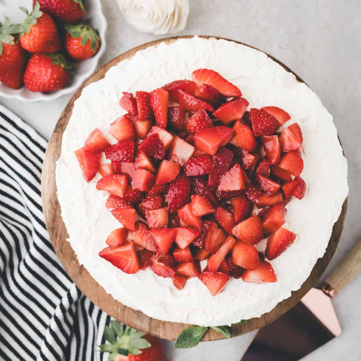 A round cake topped with whipped cream and sliced fresh strawberries sits on a wooden board. A plate of strawberries, a striped cloth, and a knife are nearby.